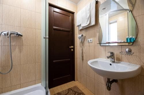 A cozy hotel bathroom features beige tiled walls, a glass-enclosed shower, a wooden door, a sink with a mirror above it, and neatly arranged toiletries, ideal for travelers seeking comfort and cleanliness.