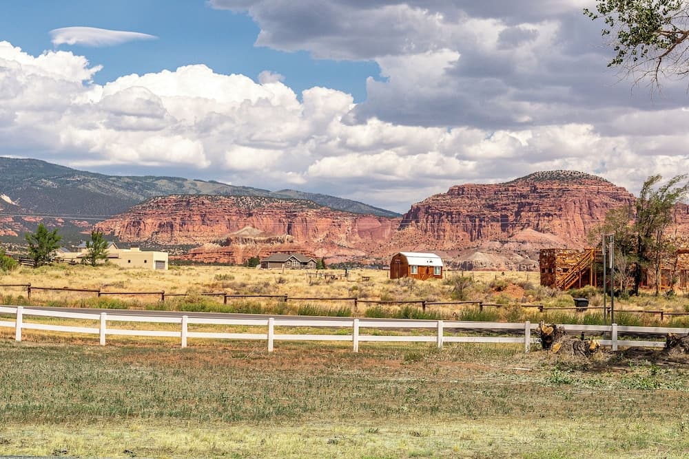 A serene rural landscape features a white fence, rustic cabins, and vibrant red rock formations under a partly cloudy sky, ideal for travelers seeking peaceful countryside retreats.