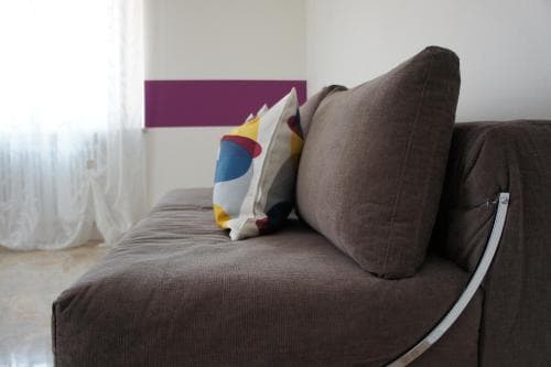 A cozy living room corner features a plush brown sofa adorned with colorful cushions, set against a minimalist backdrop with sheer curtains, ideal for travelers seeking a comfortable and stylish accommodation.