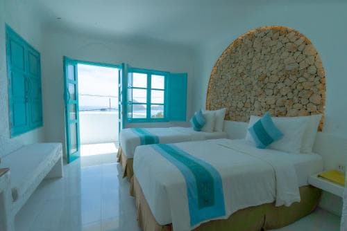 A serene hotel room with twin beds adorned in turquoise accents, a rustic stone wall feature, and open blue-framed windows inviting natural light, ideal for travelers seeking a tranquil coastal retreat.