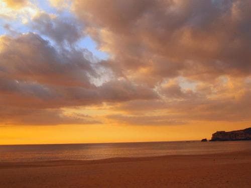 A serene beach at sunset showcases golden hues in the sky, calm waters, and distant cliffs, ideal for tranquil coastal retreats.