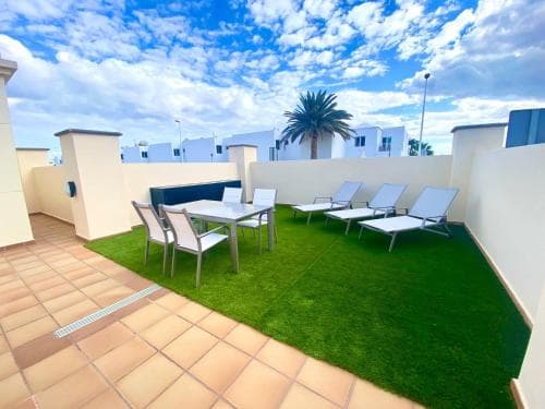 A serene rooftop terrace features artificial grass, lounge chairs, and a dining set under a vibrant blue sky, ideal for relaxing getaways in sunny destinations.
