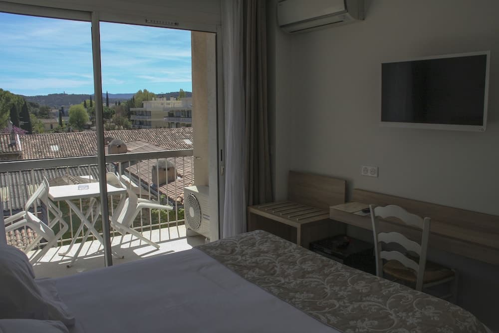 A cozy hotel room with a balcony overlooking tiled rooftops and lush greenery offers a serene retreat, featuring modern amenities like a flat-screen TV and a comfortable bed, ideal for travelers seeking relaxation and scenic views.
