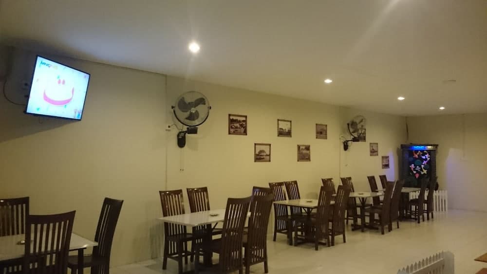 A cozy indoor dining area with wooden tables and chairs, wall-mounted fans, framed pictures, and a TV displaying colorful content, ideal for travelers seeking a relaxed meal spot.