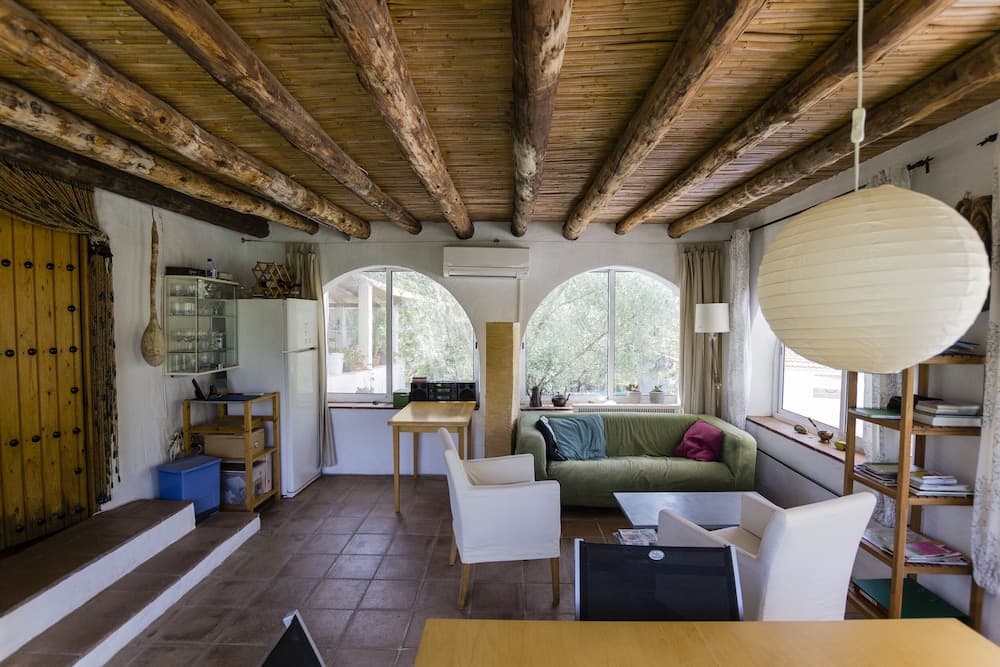 A cozy rustic living room with wooden beams, arched windows overlooking greenery, a green sofa, white chairs, and a large paper lantern creates a serene and inviting atmosphere ideal for a relaxing vacation rental.