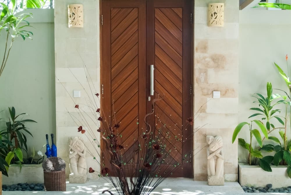 A serene tropical villa entrance features a wooden door flanked by stone carvings and lush greenery, ideal for travelers seeking a peaceful retreat.