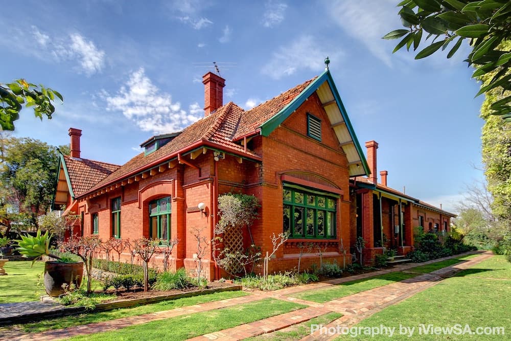 A charming red brick Victorian-style house with green-framed windows stands amidst lush greenery under a bright blue sky, ideal for travelers seeking a picturesque heritage accommodation experience.