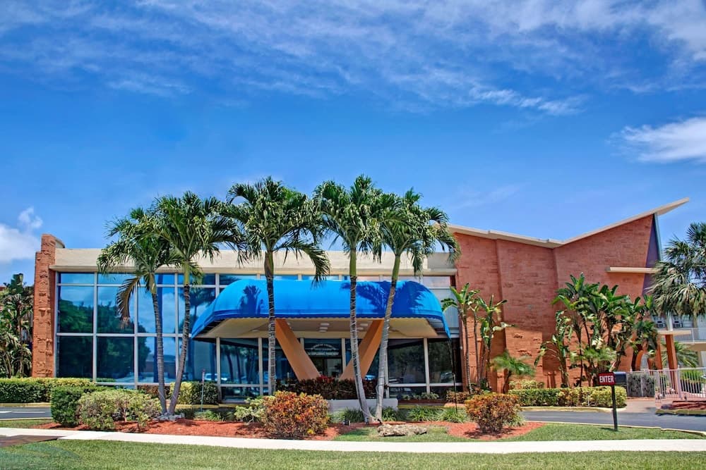 A vibrant tropical resort entrance features lush palm trees, a blue awning, and modern architecture under a clear blue sky, ideal for travelers seeking a relaxing vacation spot.