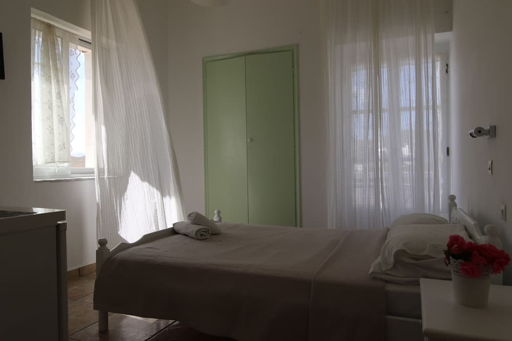 A cozy hotel room with soft natural light filtering through sheer curtains, featuring a neatly made bed, a green wardrobe, and a vase of red flowers on the bedside table, ideal for travelers seeking a serene and comfortable stay.