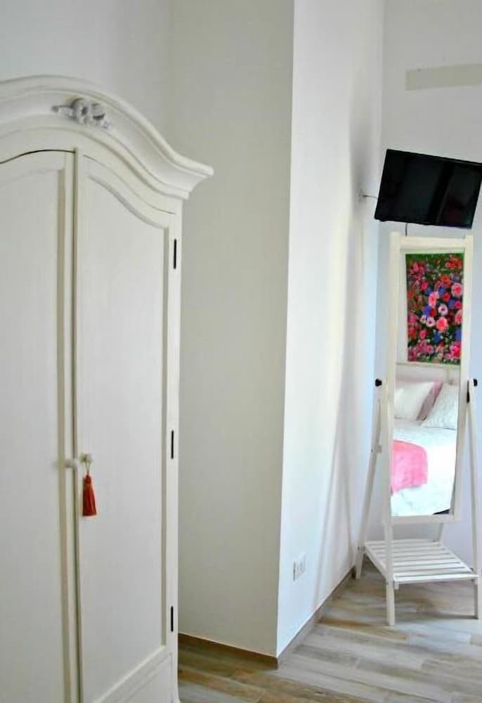 A cozy bedroom hallway features a white ornate wardrobe, a wall-mounted TV, and a floral painting reflected in a standing mirror, creating an inviting atmosphere ideal for travelers seeking a charming and comfortable stay.