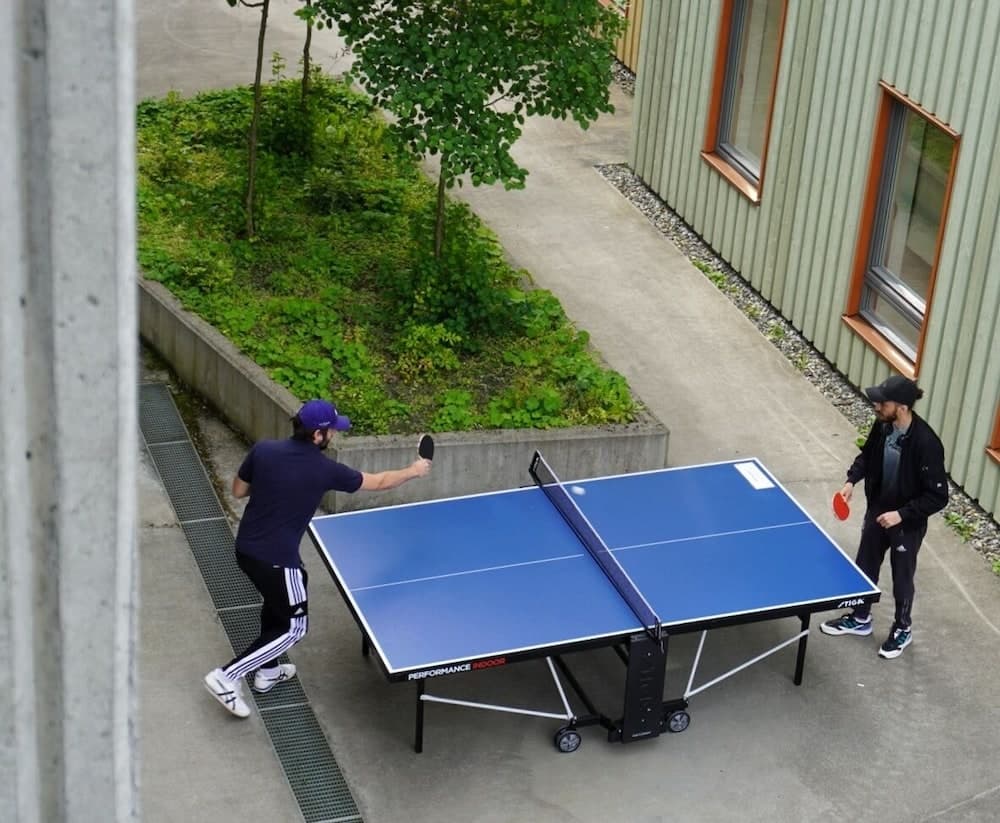 Two individuals engage in an outdoor ping pong match on a blue table surrounded by lush greenery and a modern building, creating a lively and recreational atmosphere ideal for travelers seeking active leisure activities.