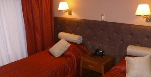 A cozy hotel room features two neatly made beds with red linens, a wooden nightstand with a phone, warm wall-mounted lamps, and curtains, ideal for travelers seeking comfort and relaxation.