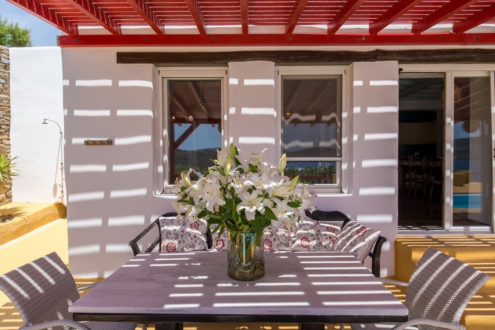 A serene outdoor patio features a cozy seating area under a red pergola, adorned with white lilies on the table, casting playful shadows on the white walls, ideal for a relaxing vacation retreat.
