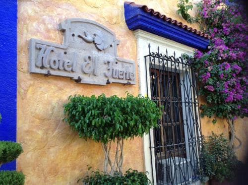 A charming hotel entrance with vibrant yellow walls, blue accents, lush greenery, and blooming flowers creates an inviting atmosphere perfect for travelers seeking a cozy and picturesque stay.