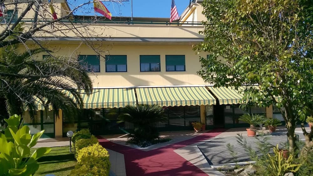 A sunny courtyard scene features a charming yellow building with striped awnings, surrounded by lush greenery and flags, ideal for travelers seeking a serene and welcoming accommodation spot.