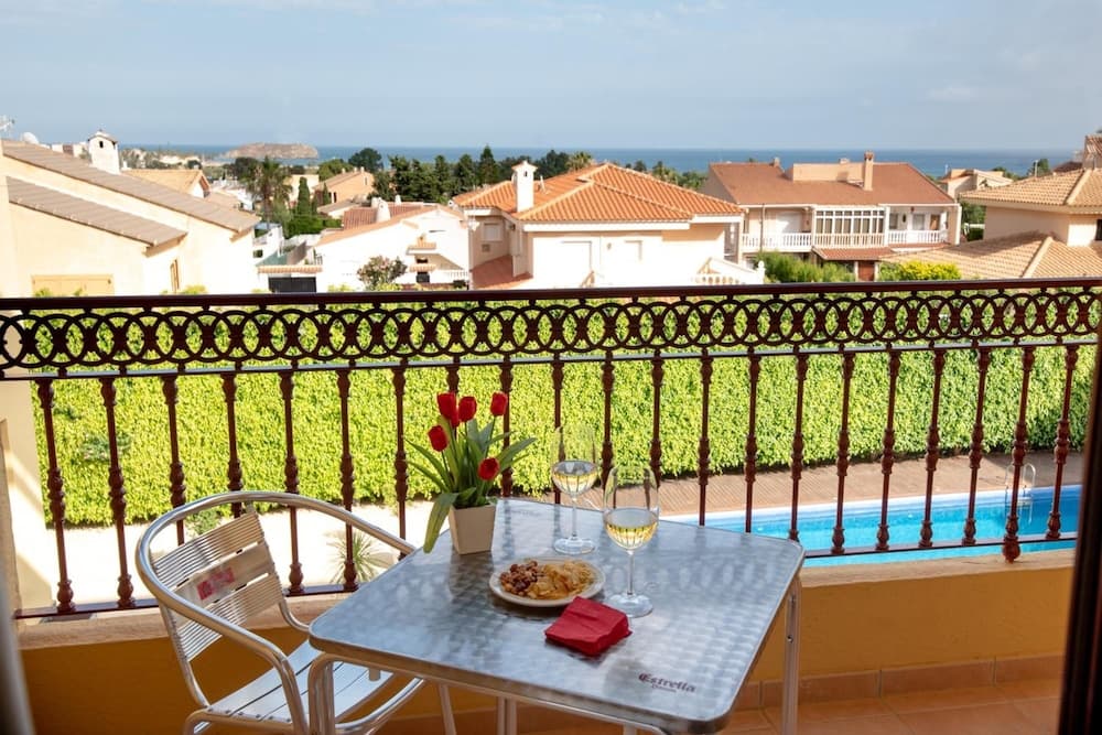 A serene balcony setup with a table adorned by tulips and wine glasses overlooks a lush hedge and a picturesque coastal town, ideal for a relaxing vacation retreat.