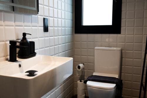 A modern bathroom with white tiled walls, a sleek sink, black fixtures, and a toilet, exudes a clean and minimalist mood, ideal for travelers seeking a stylish yet functional accommodation space.
