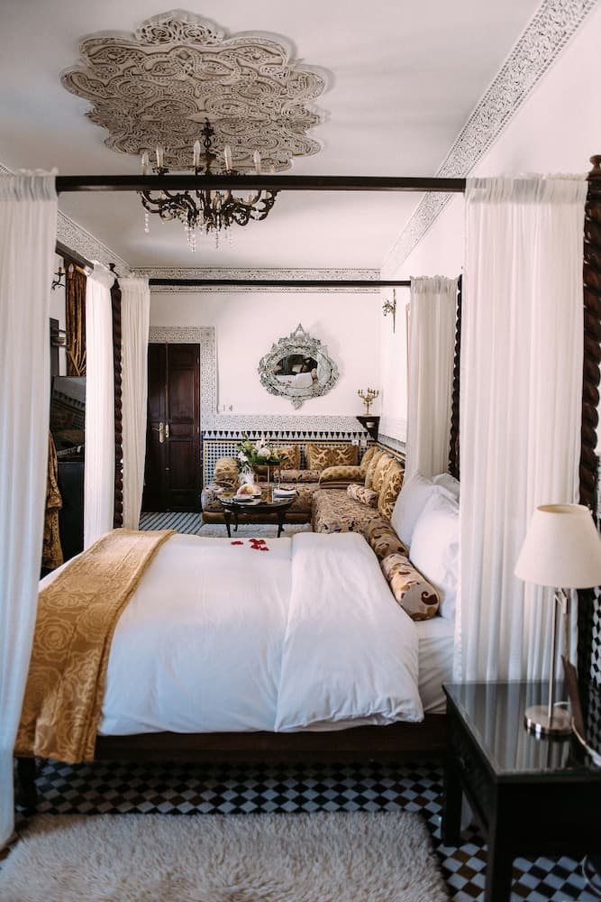 A luxurious bedroom with ornate ceiling details, a four-poster bed adorned with white linens and gold accents, and a cozy seating area featuring patterned sofas and a decorative mirror, creates an inviting and romantic atmosphere ideal for a boutique hotel stay.