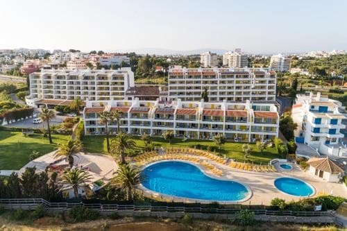 A vibrant resort scene featuring a large, inviting pool surrounded by palm trees and sun loungers, set against the backdrop of modern white buildings in a sunny coastal area, perfect for a relaxing vacation getaway.
