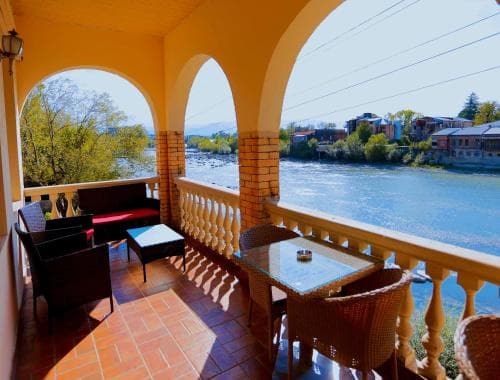 A cozy balcony with wicker furniture overlooks a serene river and quaint town under sunny skies, ideal for travelers seeking relaxation and scenic views.
