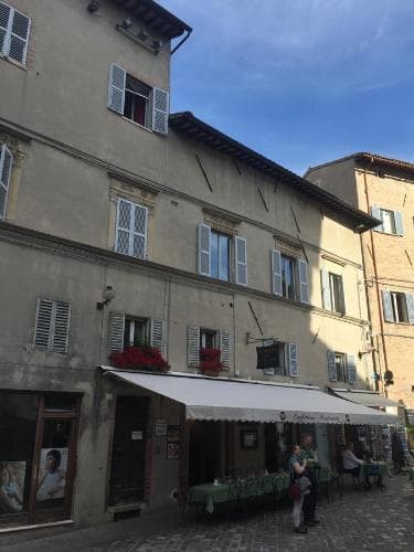A charming European street scene features a quaint café with green chairs under a striped awning, set against a historic building with shuttered windows and flower boxes, ideal for travelers seeking a cozy spot to relax and enjoy the local ambiance.