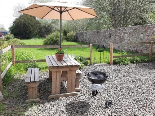 A cozy backyard patio features a wooden table under an umbrella, a barbecue grill, and a gravel ground, creating a serene outdoor dining spot ideal for relaxing getaways.