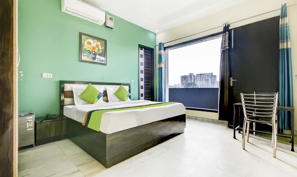 A cozy hotel room with green walls, a neatly made bed adorned with matching pillows, a balcony offering an urban view, and modern furnishings creates a welcoming atmosphere ideal for travelers seeking comfort and convenience.