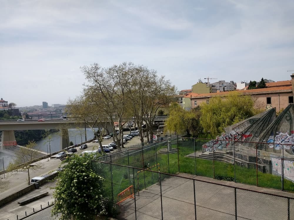 A serene riverside urban landscape features a bridge over calm waters, surrounded by greenery, graffiti-covered walls, and residential buildings, ideal for travelers seeking a blend of nature and city exploration.