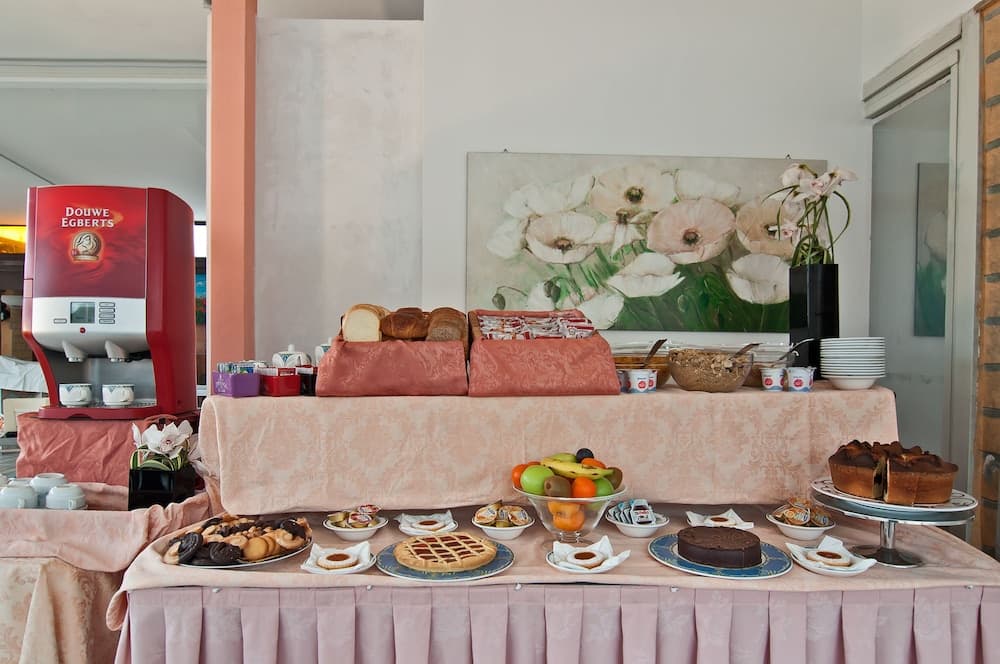 A cozy hotel breakfast buffet features an inviting spread of pastries, fruits, cakes, and coffee, set against a soft pink tablecloth and floral artwork, perfect for travelers seeking a warm morning start.