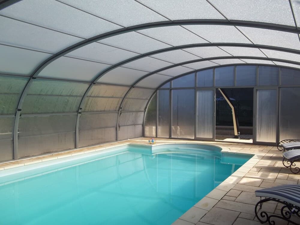 A serene indoor pool area under a translucent arched roof features calm turquoise water, lounge chairs, and glass doors, ideal for year-round relaxation and leisure travel.
