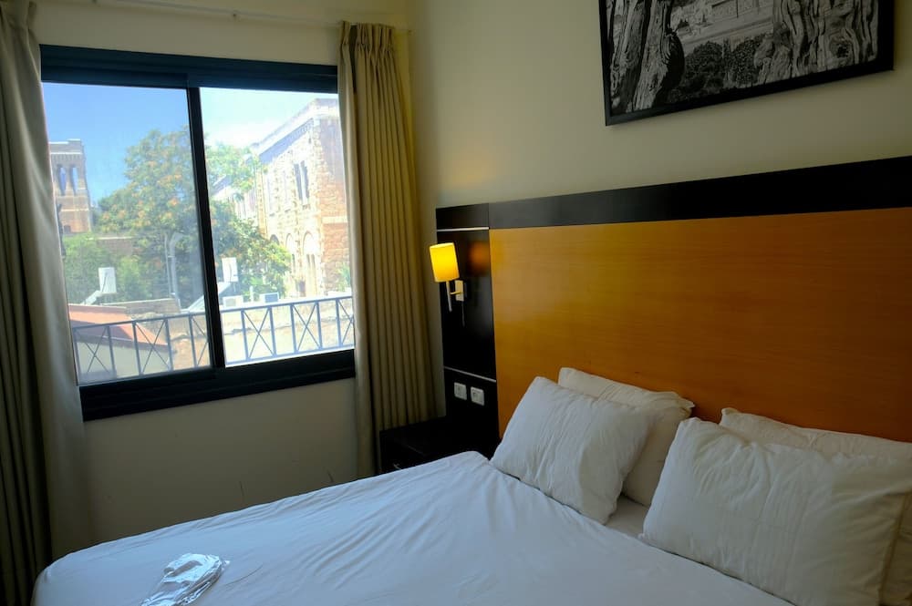A cozy hotel room with a neatly made bed, wooden headboard, and warm lighting offers a serene view of historic architecture through its large window, ideal for travelers seeking comfort and cultural ambiance.
