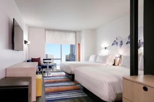A modern hotel room with two cozy beds, a vibrant striped rug, and a cityscape view through large windows, ideal for travelers seeking comfort and urban proximity.