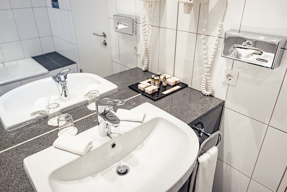 A modern hotel bathroom features sleek white sinks, a granite countertop, neatly arranged toiletries, and a clean, minimalist design ideal for travelers seeking comfort and convenience.