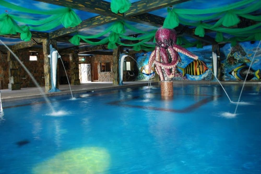 An indoor aquatic playground features a vibrant octopus fountain surrounded by colorful fish murals, green draped ceilings, and playful water jets, creating a whimsical and inviting atmosphere ideal for family-friendly vacations.