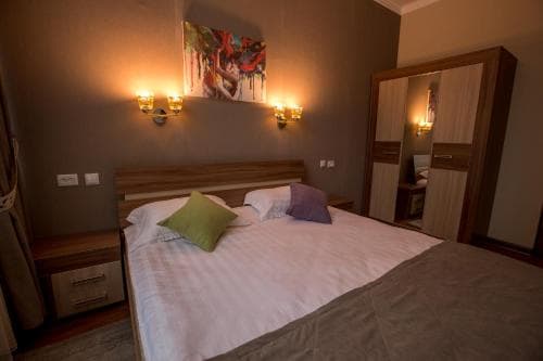A cozy hotel room features a neatly made bed with colorful pillows, warm wall sconces, a vibrant abstract painting, and a mirrored wardrobe, ideal for travelers seeking comfort and style.