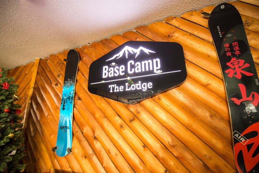 A cozy ski lodge entrance features a wooden wall adorned with "Base Camp The Lodge" signage, flanked by colorful snowboards and a festive Christmas tree, ideal for winter sports enthusiasts seeking a warm retreat.