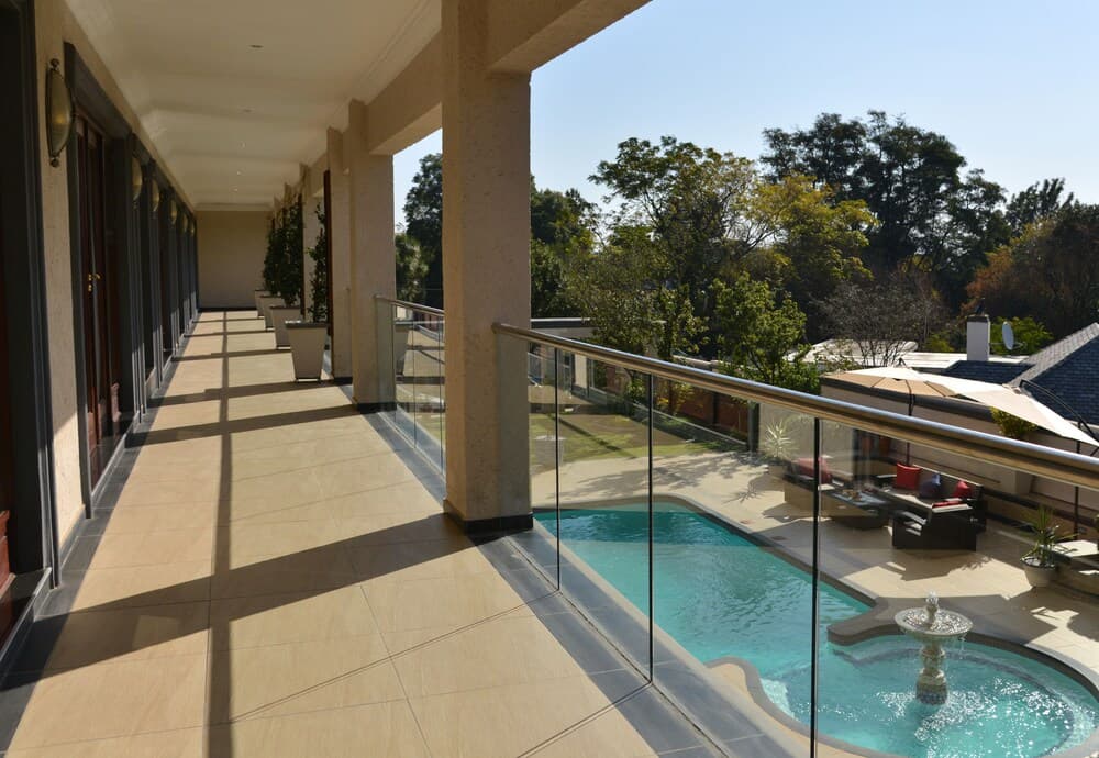 A serene luxury villa balcony overlooks a tranquil pool area surrounded by lush greenery, featuring glass railings, elegant columns, and cozy seating, perfect for a relaxing vacation retreat.