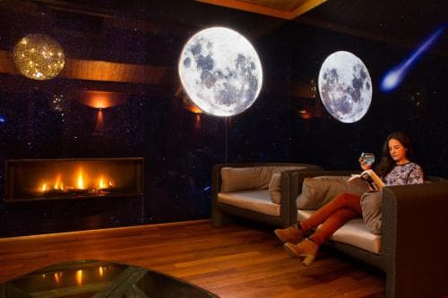 A serene indoor lounge features a cozy fireplace, celestial moonlight projections on the wall, and a relaxed woman enjoying a drink, ideal for a tranquil getaway or romantic evening.
