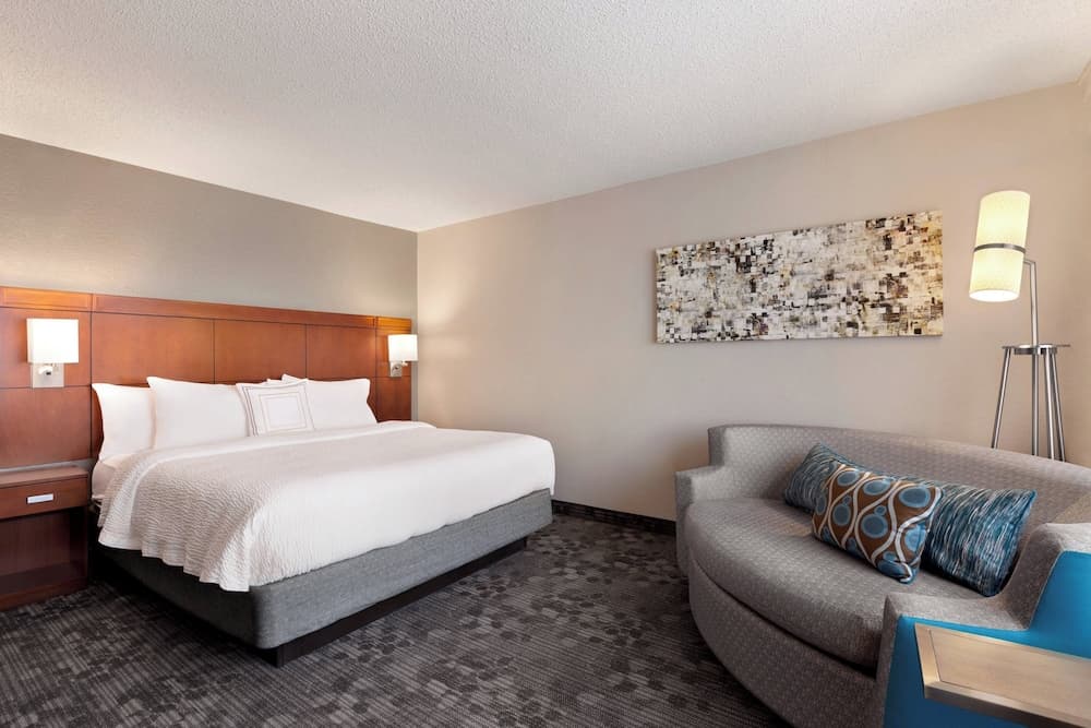 A cozy hotel room features a neatly made bed with white linens, a modern sofa adorned with patterned pillows, and ambient lighting, ideal for travelers seeking comfort and relaxation.