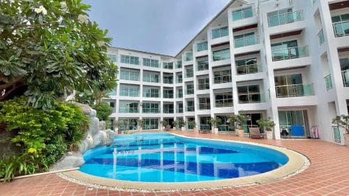 A serene courtyard features a vibrant blue pool surrounded by lush greenery and modern multi-story apartments, ideal for a relaxing vacation stay.