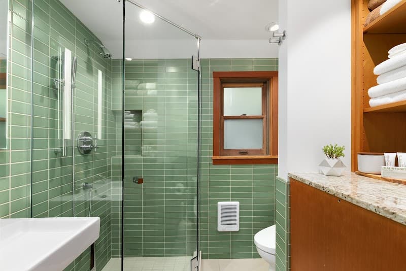 A serene coastal cabin bathroom features green tiled walls, a modern glass shower, a small window with a wooden frame, and neatly arranged towels on a wooden shelf, ideal for travelers seeking a relaxing seaside retreat.