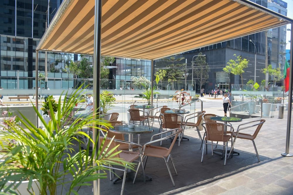 A serene urban café patio shaded by a striped awning features cozy seating, lush potted plants, and a view of modern glass buildings, ideal for travelers seeking a relaxing spot to unwind in a bustling city.