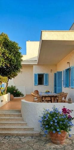 A serene Mediterranean patio features a cozy dining area under a clear blue sky, surrounded by vibrant flowers and lush greenery, ideal for travelers seeking a tranquil vacation retreat.