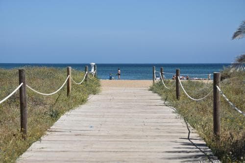 A serene wooden boardwalk lined with rope railings leads to a tranquil beach under a clear blue sky, ideal for a peaceful coastal getaway.