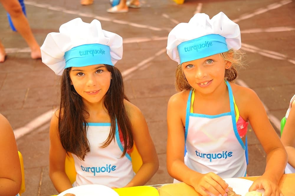 Two cheerful girls wearing turquoise-themed chef hats and aprons enjoy a culinary activity at an outdoor event, perfect for family-friendly travel experiences.