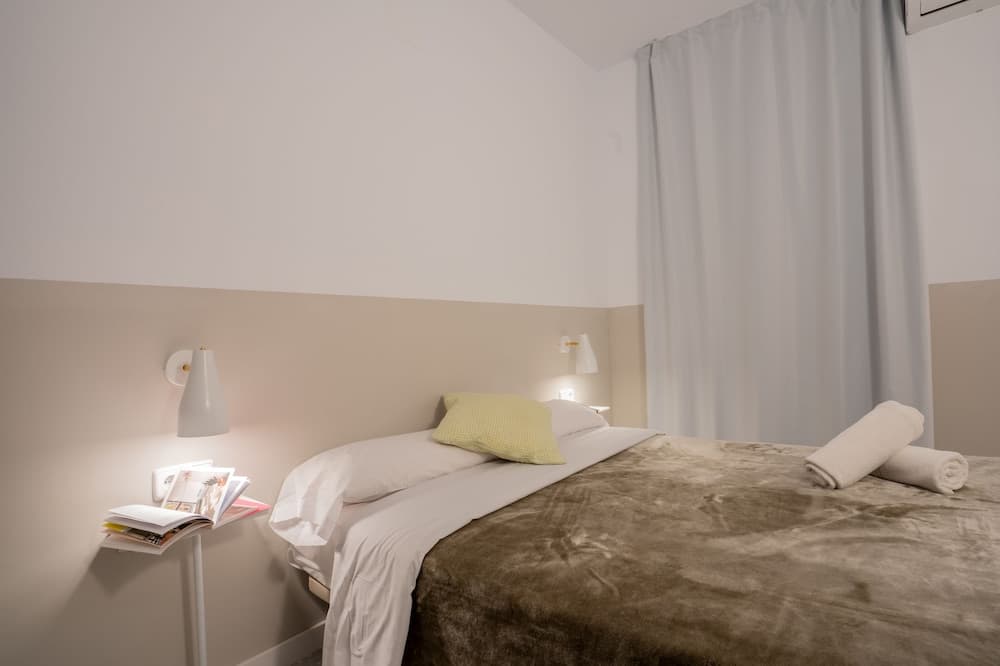 A cozy hotel room features a neatly made bed with white linens, a yellow pillow, rolled towels, soft lighting from wall-mounted lamps, and a side table with magazines, ideal for travelers seeking comfort and relaxation.
