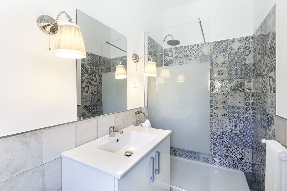 A modern bathroom features a sleek white vanity, patterned blue tiles, a glass shower enclosure, and soft lighting from wall-mounted lamps, creating a serene and stylish space ideal for travelers seeking comfort and design in their accommodations.