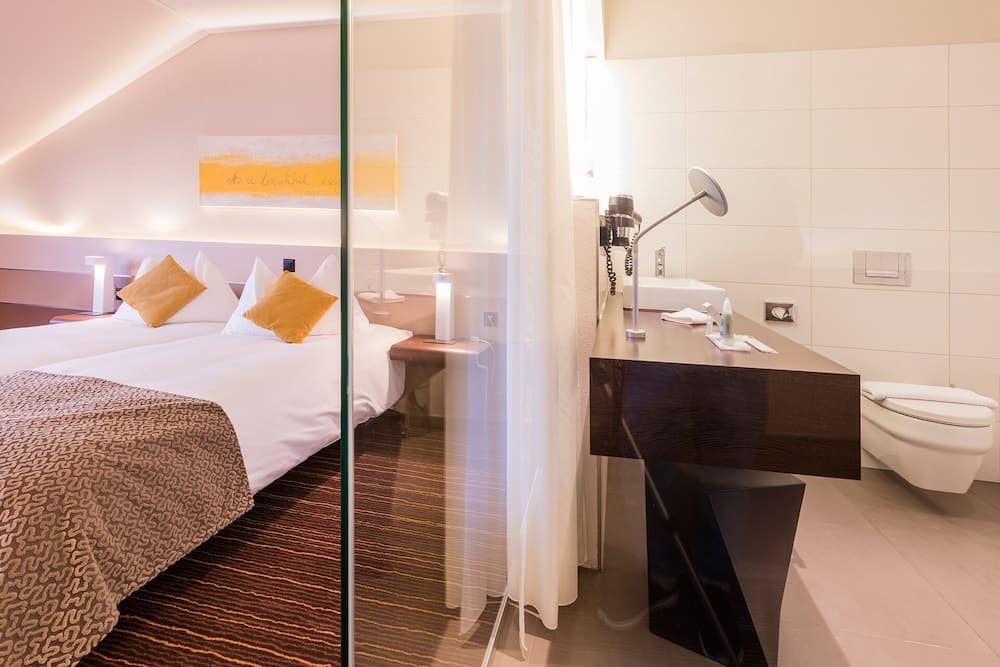 A cozy hotel room with a neatly made bed adorned with orange pillows, a sleek desk with a modern lamp, and an open bathroom featuring white tiles, ideal for travelers seeking comfort and style.