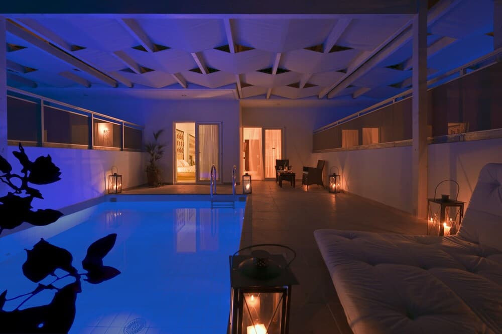 A serene indoor pool area bathed in soft blue light, featuring candle lanterns, plush seating, and a geometric ceiling, ideal for relaxation and luxury retreats.