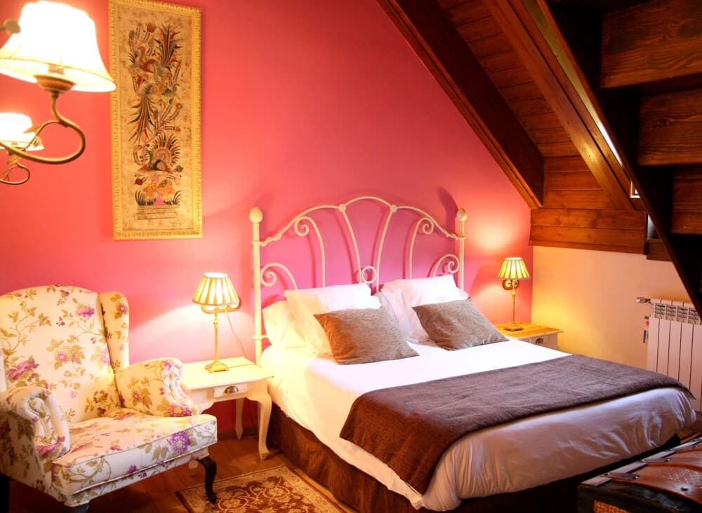 A cozy attic bedroom with vibrant pink walls, a charming floral armchair, and warm lighting creates an inviting atmosphere ideal for a romantic getaway.